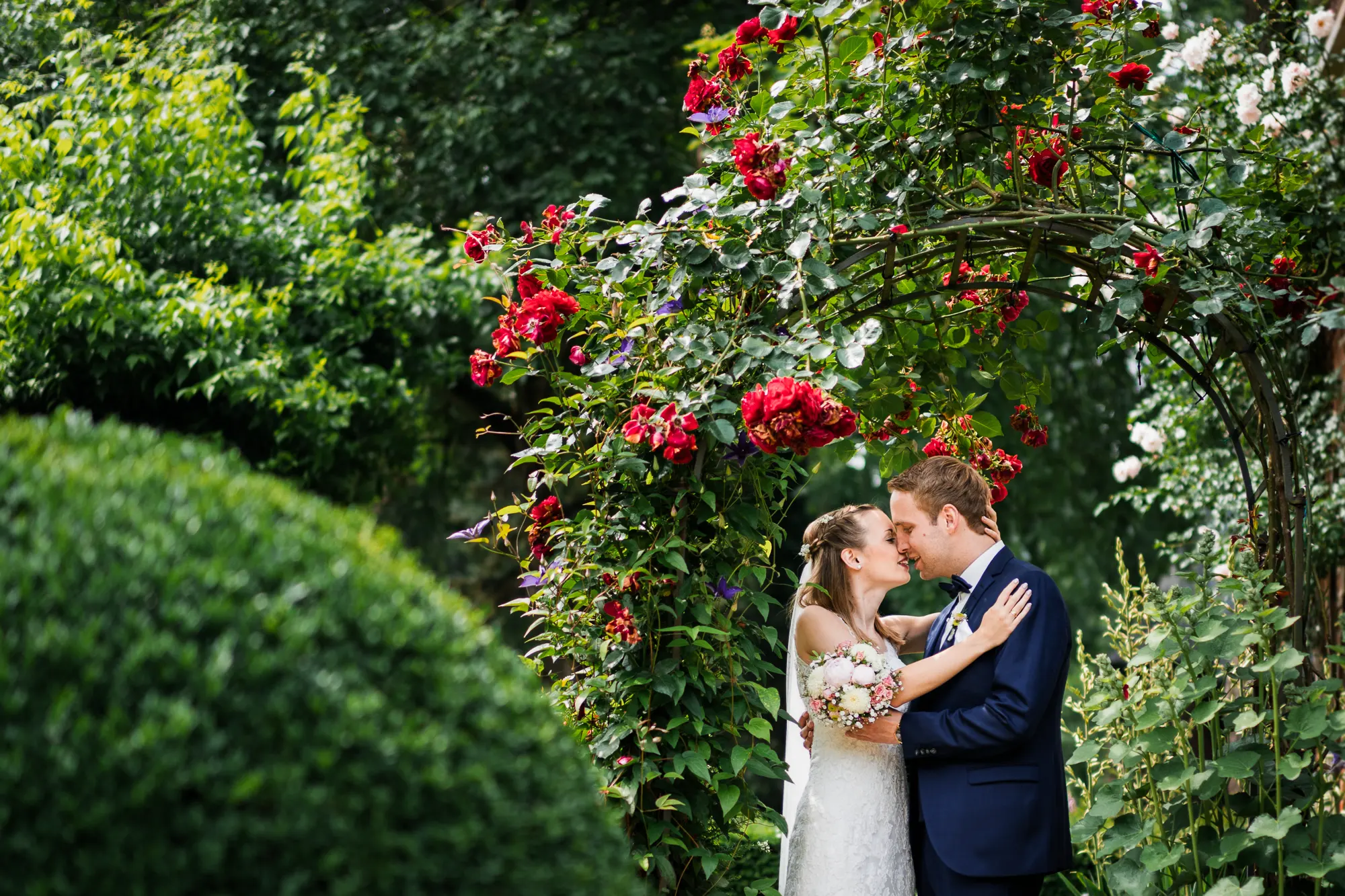 Am Hochzeitstag küsst sich ein Brautpaar unter einem blühenden Rosenbogen im Garten des Haus Welbergen