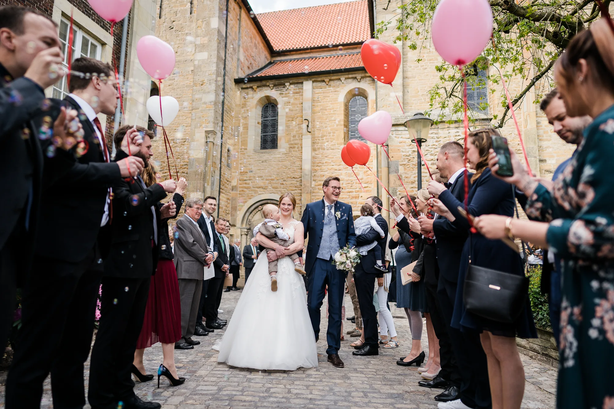 Brautpaar zieht mit ihrem Kind auf dem Arm durch einen Spalier aus Luftballons und Seifenblasen vor der St. Johannes Baptist Kirche in Langenhorst - Hochzeitsfotograf Ochtrup
