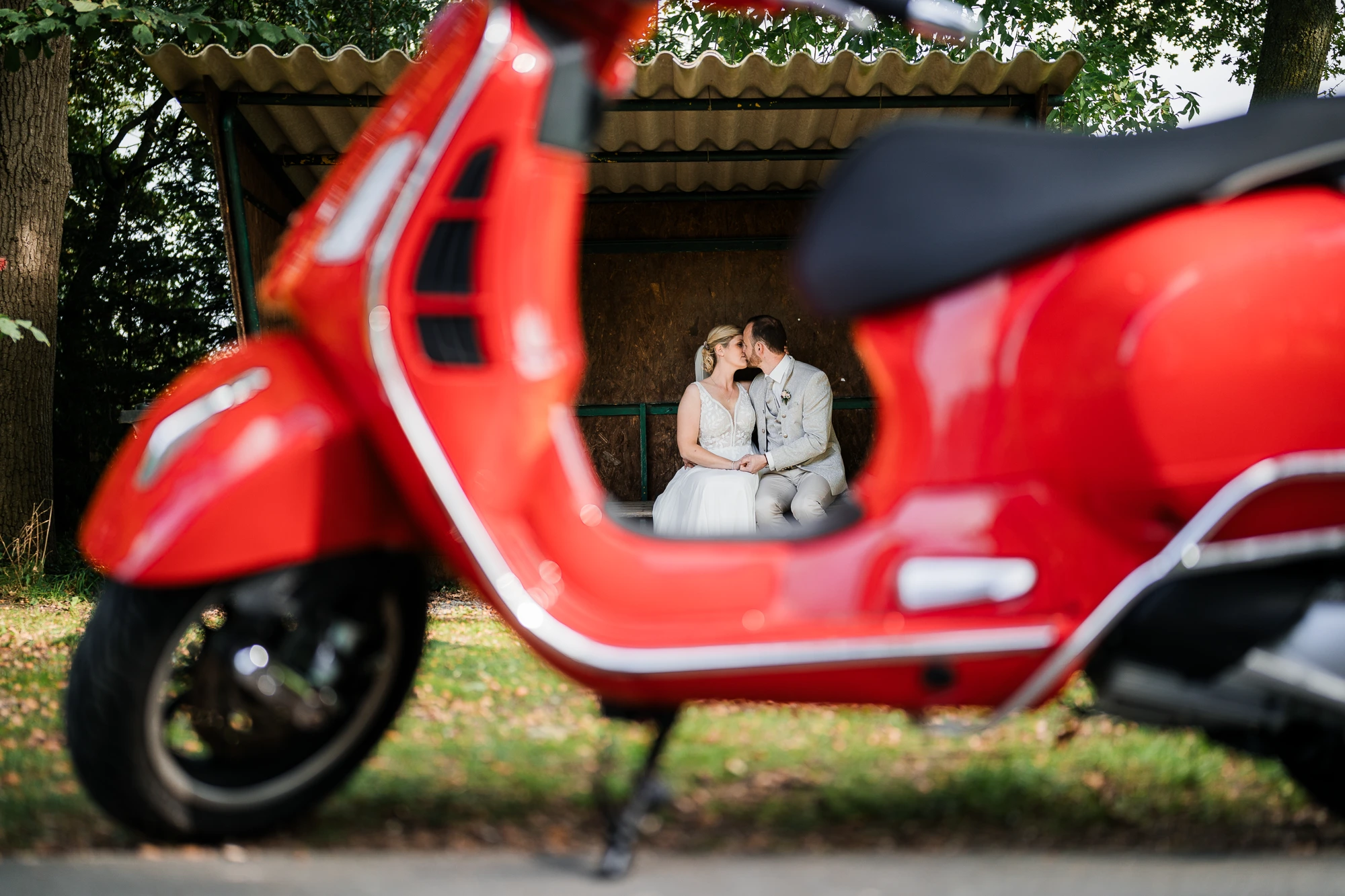 Brautpaar küsst sich an einer Bushaltestelle in der Ochtruper Wester, gerahmt durch eine rote Vespa im Vordergrund